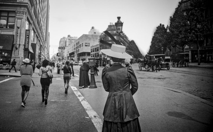 Femme en robe ancienne observant une rue animée, avec d'autres piétons et des bâtiments.