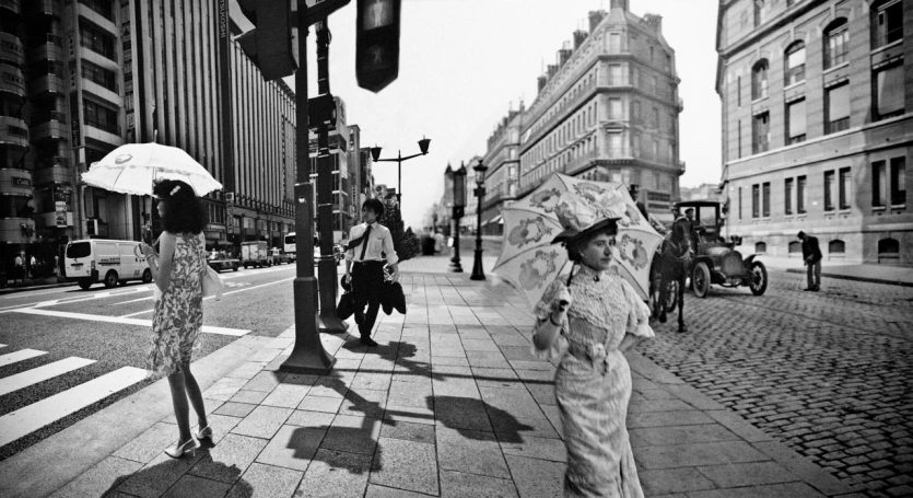 Deux femmes avec des parasols marchent dans une rue animée en noir et blanc.